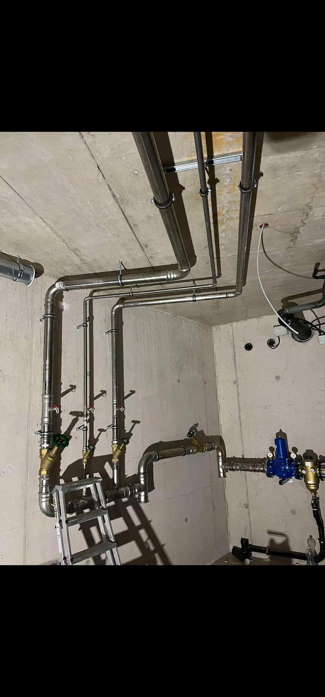 Az Ösküben található HydroArt Kft. Épületgépészet területén végzett munkáját bemutató képen a mennyezetből induló rozsdamentes acél csövek láthatók, amelyek különböző irányokba kanyarodnak. A csövek között szelepek és egy mérőberendezés is látható, amely a precíz működést segíti. A háttérben egy létra is található, ami a telepítési munkákhoz szükséges.