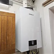 Fehér kazán látható egy falra szerelve, amely a HydroArt Kft. épületgépészeti projektjének része Öskü városában. A kazán modern kialakítása és a falon lévő csövek jól mutatják az épületgépészet területén végzett precíz munkát. Az elegáns design és a funkcionális elhelyezés tükrözi a cég szakértelmét az épületgépészetben.