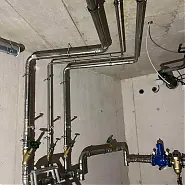Az Ösküben található HydroArt Kft. épületgépészeti munkáit bemutató képen rozsdamentes acél csövek láthatók, amelyek különböző irányokba kanyarodnak. A csövekhez szelepek és csatlakozók tartoznak, amelyek az épületgépészet szakszerű kivitelezését jelzik. A háttérben a betonfalak és a szerelési elemek is jól láthatók, ami a precíziós munkát tükrözi.