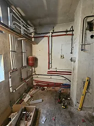 A képen egy épületgépészeti munka látható, ahol a falon piros és fehér csövek futnak, valamint egy vízmelegítő tartály található. Az Ösküben található HydroArt Kft. által végzett munkálatok során a helyiségben látható állapot még befejezés előtt áll. A padlón különféle szerszámok és anyagok hevernek, ami az építkezés aktív fázisára utal.