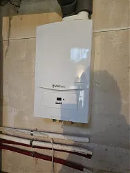 A képen egy modern fali kazán látható, amely a HydroArt Kft. épületgépészeti munkájának részeként került telepítésre Öskün. A kazán fehér színű, kompakt kialakítású, és a falon helyezkedik el. A készülék alatt piros és fehér csövek láthatók, amelyek az épületgépészeti rendszerek részei.