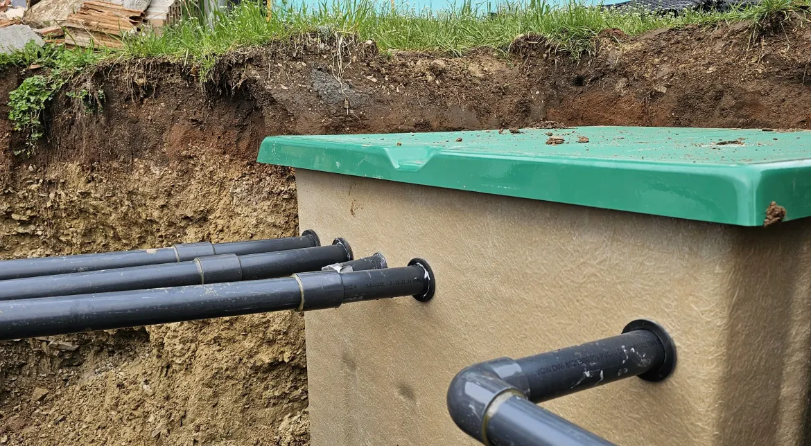 A képen egy frissen ásott árkot látunk Ösküben, ahol a HydroArt Kft. épületgépészeti munkálatai zajlanak. Az árkot a földbe süllyesztett zöld fedéllel ellátott tartály és több fekete csőcsonk jellemzi, amelyek az épületgépészethez szükséges víz- vagy csatornarendszert szolgálják. A környezet zöld fűvel és néhány építkezési anyaggal van körülvéve.