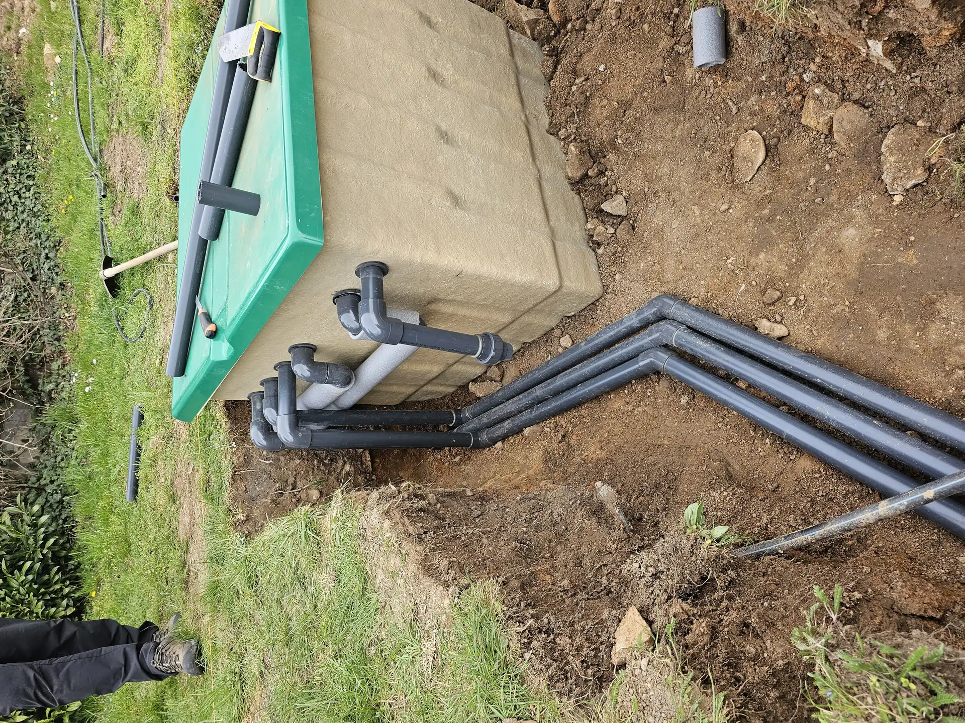 A képen egy épületgépészeti munkálat látható Öskü területén, ahol a HydroArt Kft. szakemberei csöveket telepítenek. A földbe ásott árokban fekvő fekete csövek különböző irányokba futnak, míg egy zöld fedelű tartály is jól látható a háttérben. Az építkezés körüli terület zöld fűvel borított, ami a munkálatok természetes környezetét mutatja.
