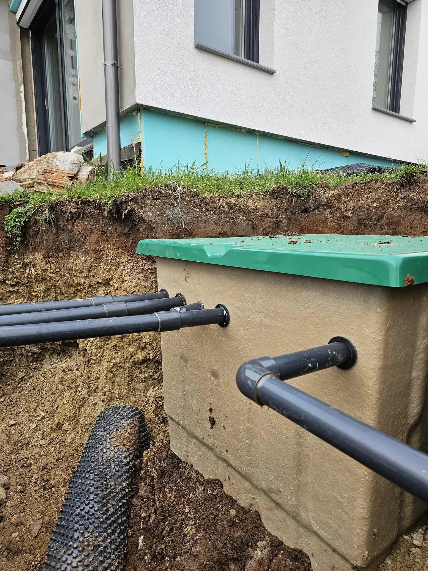 Az Ösküben található HydroArt Kft. épületgépészeti munkálatai során készült képen egy frissen telepített szennyvízcsatorna látható, amely beton alapra van helyezve. A kép előterében fekete PVC csövek futnak, míg a háttérben a ház falának részletei és a zöld szigetelés is megfigyelhető. A terület rendezése és a csövek elhelyezése az épületgépészet szakszerű kivitelezését tükrözi.