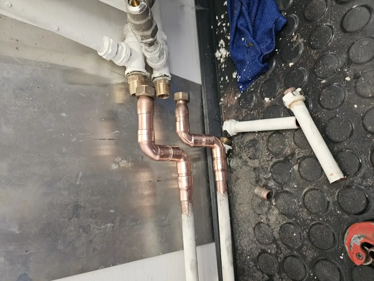 Az Ösküben található HydroArt Kft. épületgépészeti munkálatait bemutató képen réz és műanyag csövek láthatók, amelyek szakszerűen vannak összekötve. Az installáció a fal mellett helyezkedik el, és láthatóan precíz munkavégzést tükröz. A képen a csöveken kívül néhány szerszám és egy kék rongy is található, amely a munkaterület tisztaságát hangsúlyozza.