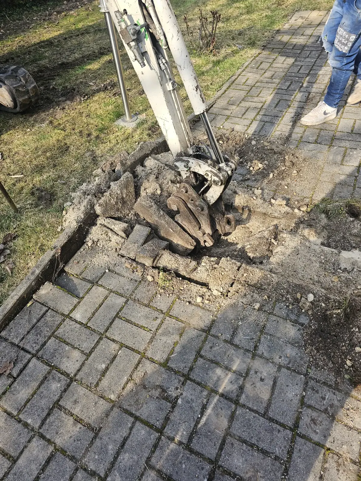 Az Ösküben található HydroArt Kft. Épületgépészet területén végzett munkáját mutatja be az image slot: gallery_06. A képen egy gép látható, amely éppen egy térkőből készült járdát bont le, a környezetben fű és föld látható. A munka során a gép a járda köveit és az alatta lévő anyagokat távolítja el.