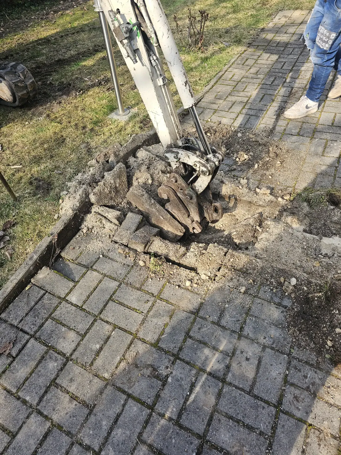 A képen egy épületgépészeti munkálat látható, ahol egy gép ásási folyamatot végez egy térkővel burkolt járdán Öskü városában. A gép körül törmelék és föld található, míg a háttérben egy zöld fűsáv látható. A HydroArt Kft. szakemberei éppen dolgoznak a területen, hogy biztosítsák az épületgépészeti rendszer megfelelő telepítését.