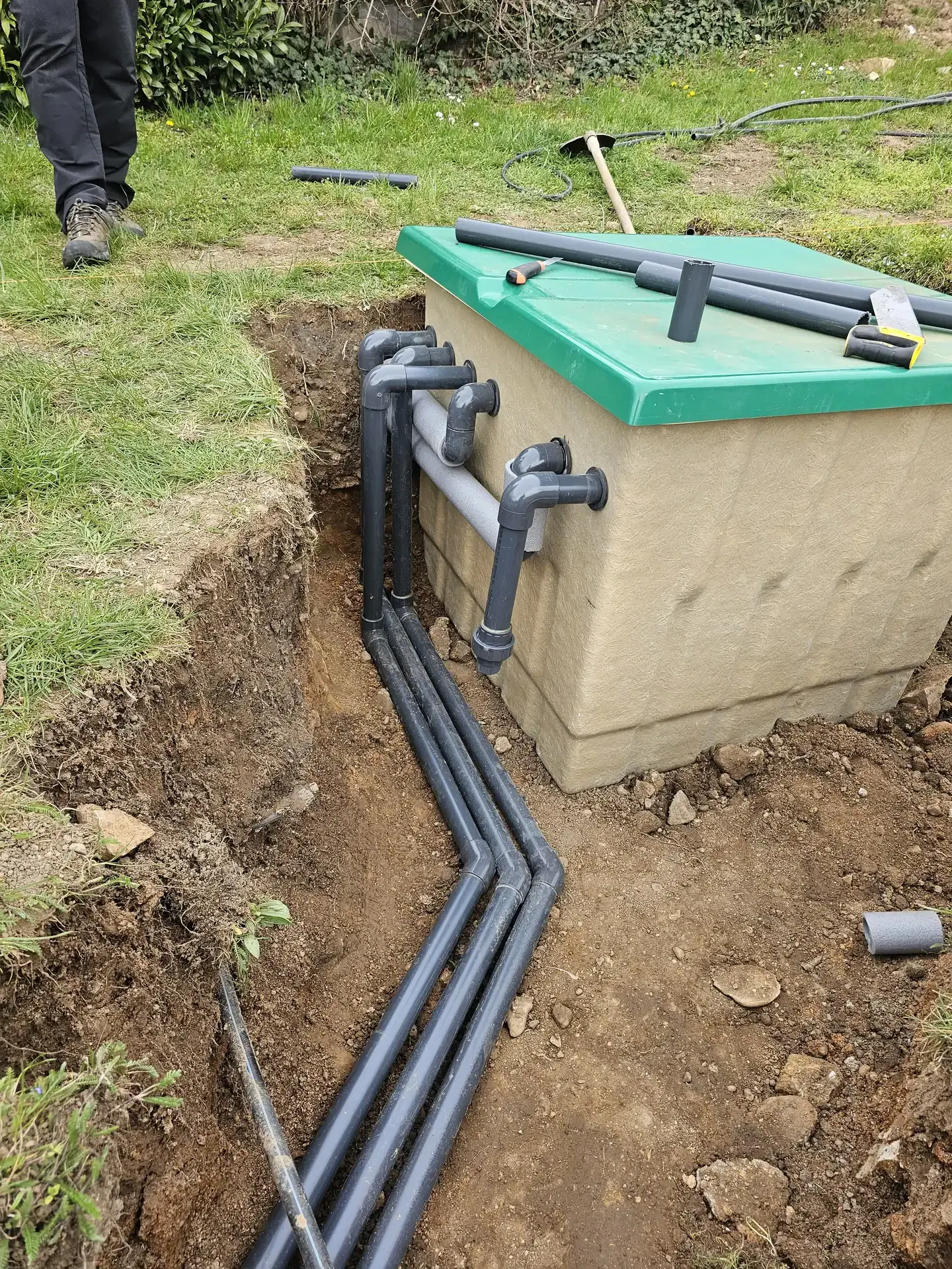 Az Ösküben található HydroArt Kft. Épületgépészet munkálatai során készült kép, amelyen egy földbe ásott tartály látható zöld fedéllel. A tartályból több fekete cső vezet ki, amelyek a talajban futnak. A környező terület füves, és a háttérben látható néhány szerszám is, amelyek az épületgépészeti munkához kapcsolódnak.