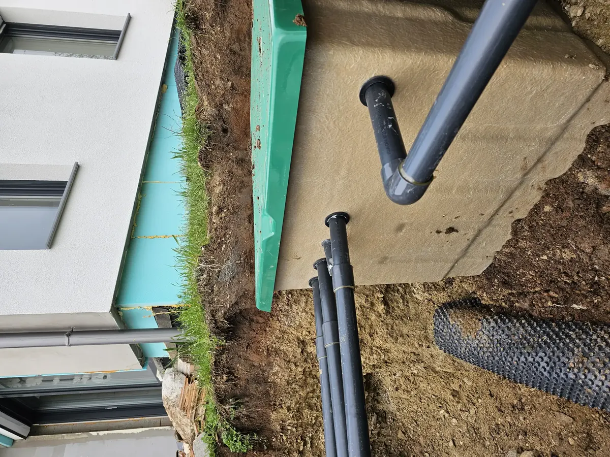 Az Ösküben található HydroArt Kft. épületgépészeti munkálatai során készült képen egy frissen kiásott terület látható, ahol szürke PVC csövek futnak a földbe, valamint egy beton alapú tartály részletei is megjelennek. A háttérben modern épület falai és zöld szigetelés látható, amely a vízszigetelés része. Az épületgépészet precíz munkavégzését tükrözi.