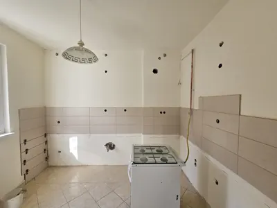 Az Ösküben található HydroArt Kft. Épületgépészet projektjének képe, amely egy felújítás alatt álló konyhát ábrázol. A falakat világos csempe borítja, és a fehér falon láthatók a villanyszerelés nyomai. A konyhában egy modern gáztűzhely helyezkedik el, amely készen áll a beépítésre. A világító lámpa elegáns formája kiemeli a tér letisztult vonalait.